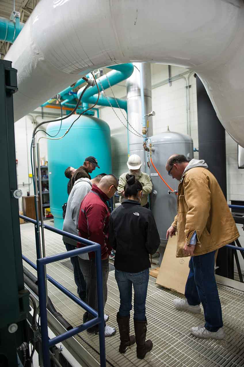 Photo of a group of people looking down at the grated floor of a heating area inside a building.