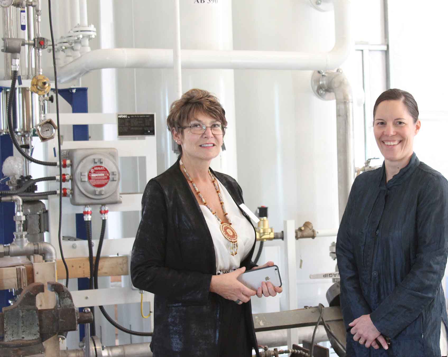 Photo of two women standing in front of a series of pipes in an indoor heating facility.