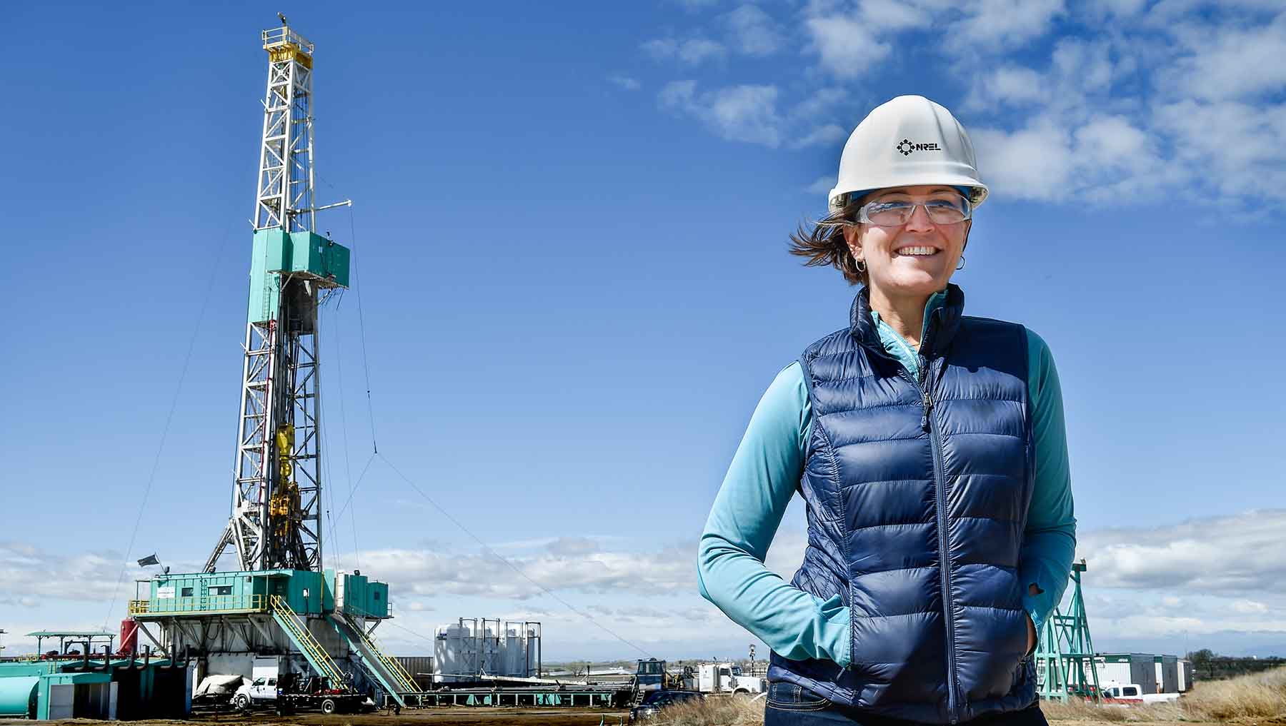 A woman wearing a hard hat stands in a field near an oil and gas well.