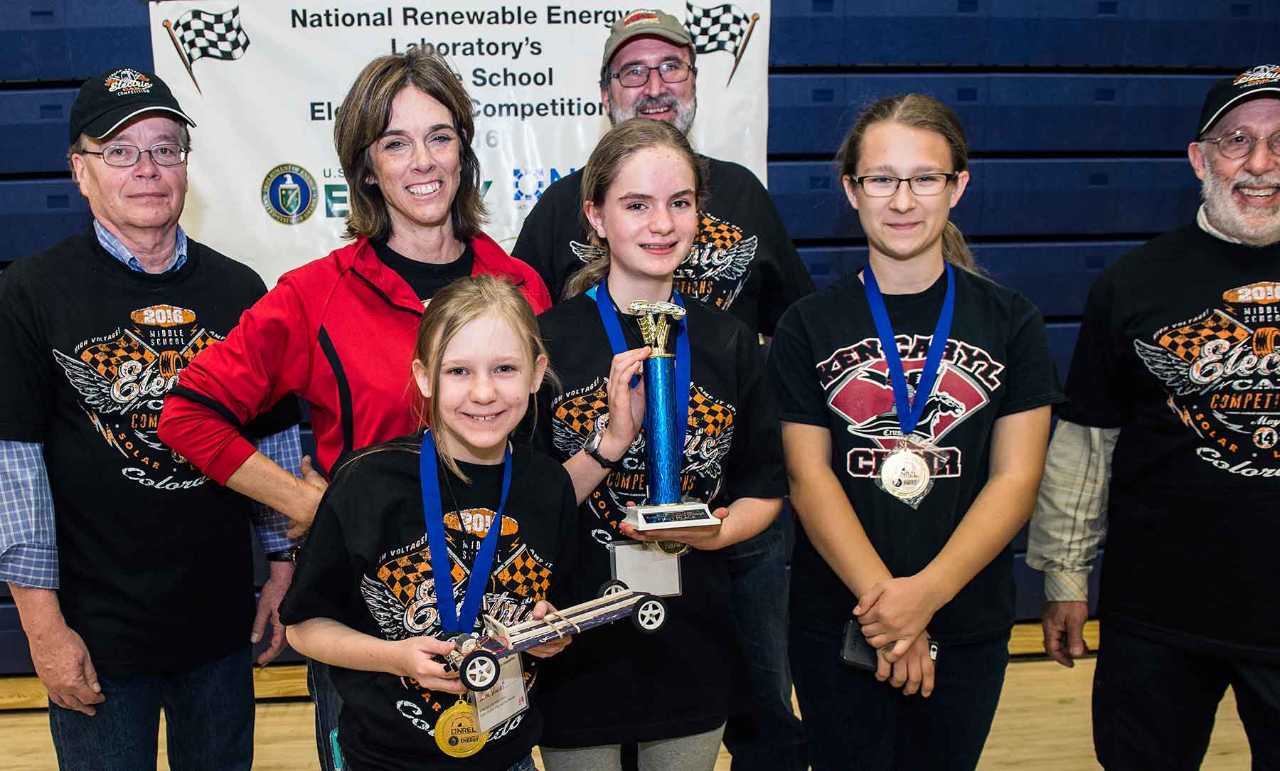 Three young girls smile, holding trophies and a model car, as four adults stand behind them in a gym.