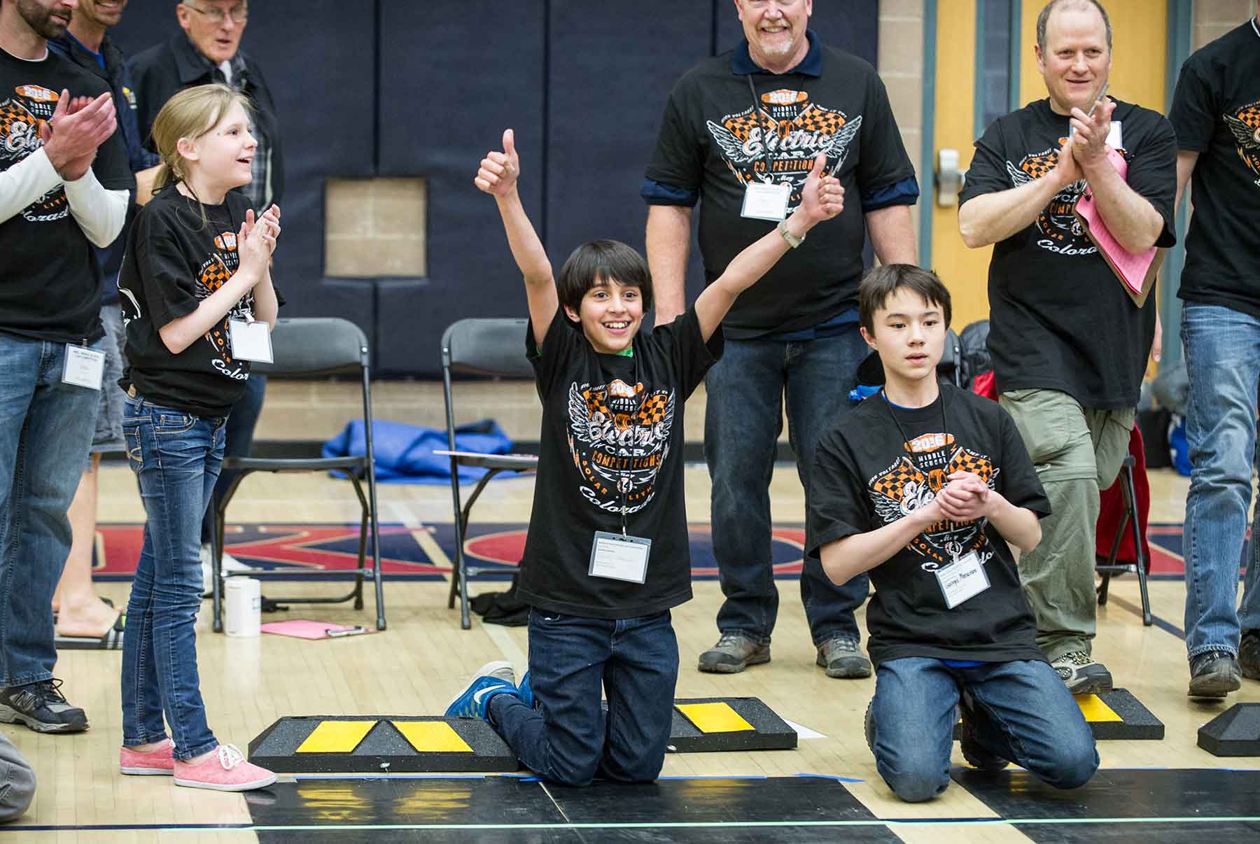 Two kneeling boys and a standing, smiling girl look down a racetrack in a gym.