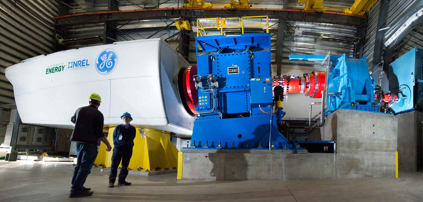 Two men in hardhats and safety glasses stand in front of equipment having a conversation. The blue and white dynamometer equipment has a white wind turbine drivetrain attached to it for testing.