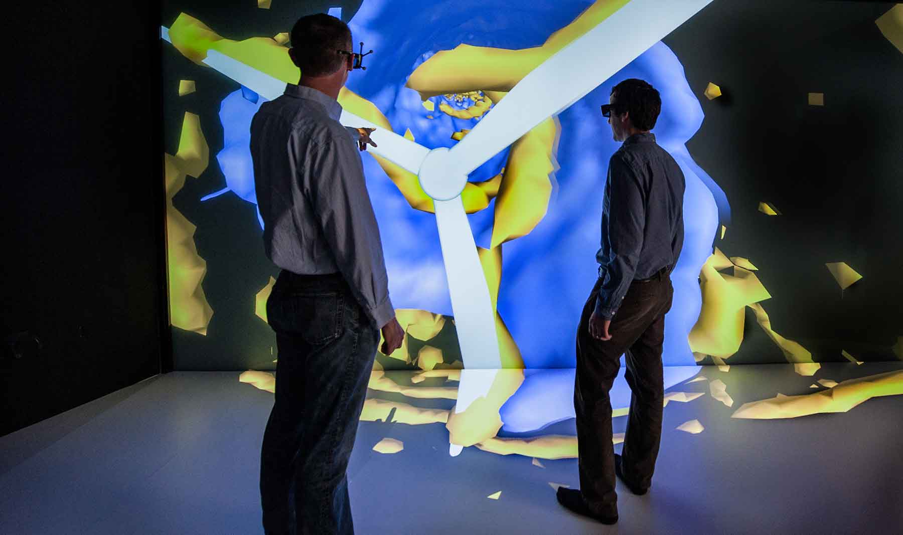 An engineer wearing special glasses points to an image on a large visualization as another engineer looks on.