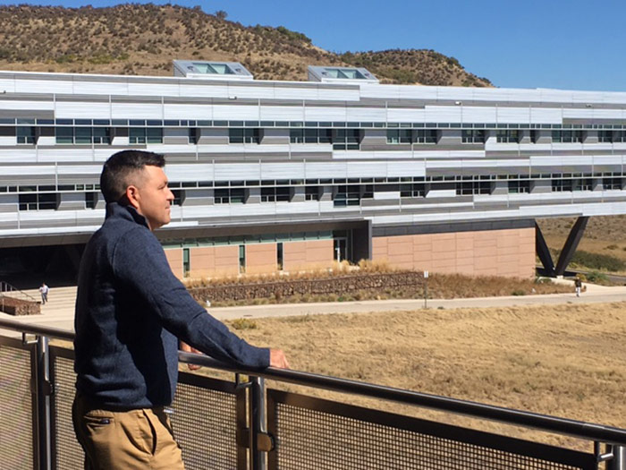 A man on a balcony looks out at a field with a building behind him.