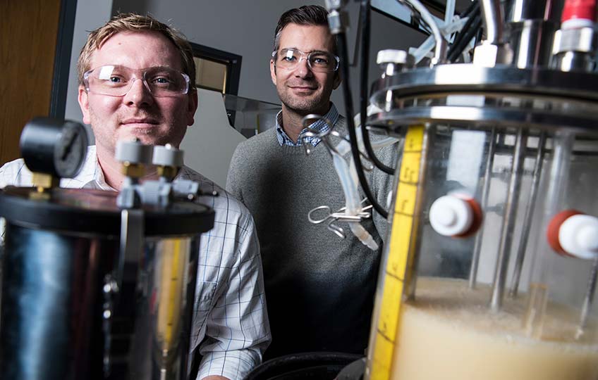 Two men stand next to a container of a murky liquid.