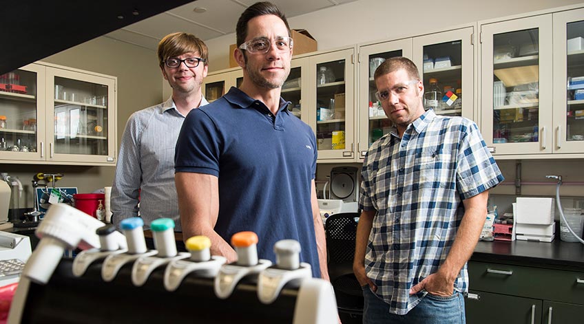 Photo shows three men standing in a laboratory.