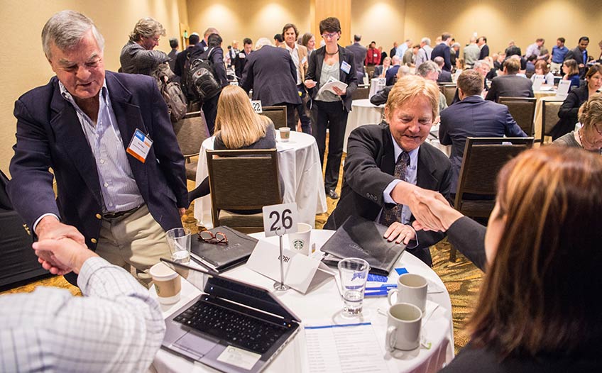 Two men shake hands with a man and woman seated at a table in a room filled with people in similar meetings at similar tables.