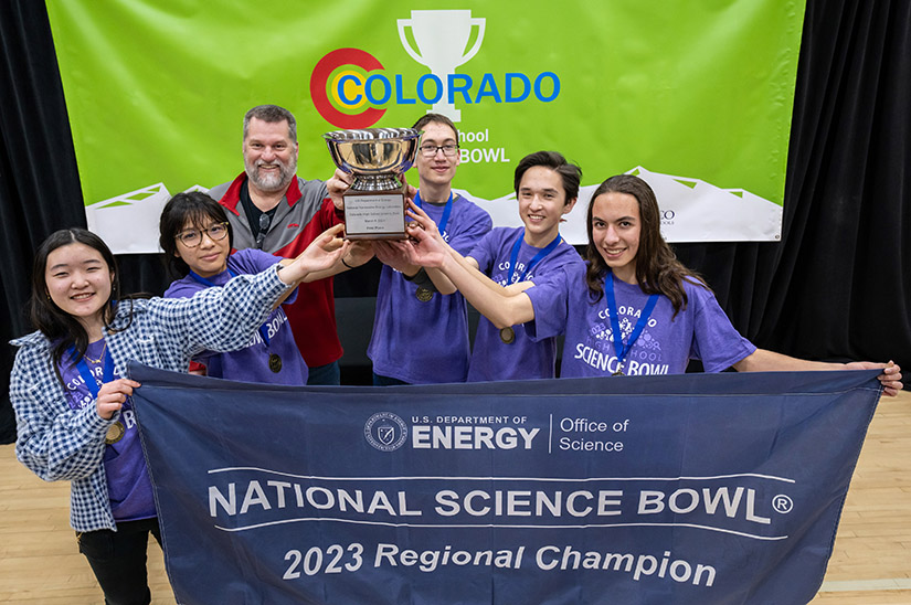 Five smiling students and a teacher holding a trophy and banner.