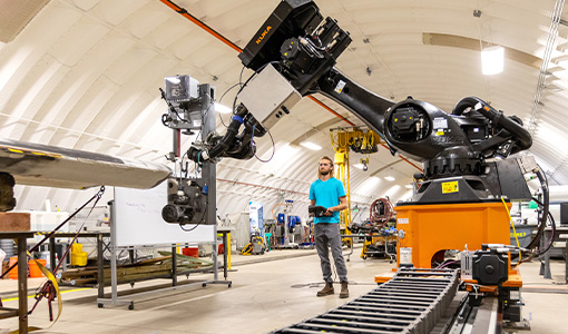 A person operates a robot arm next to a turbine blade in a large laboratory.