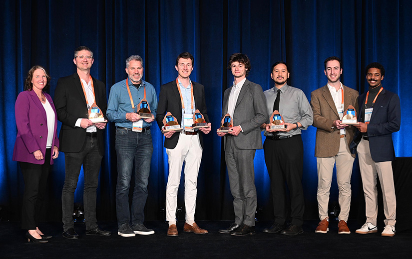 Eight people stand on a stage while holding awards.