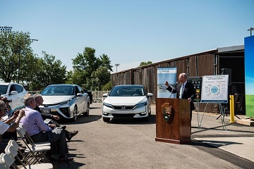 Photo of a man standing outside at a podium and giving a speech to a seated crowd.