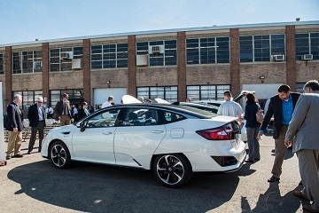 Photo of a white four-door vehicle parked in a lot next to two other vehicles. Several people are standing and walking around the vehicles.