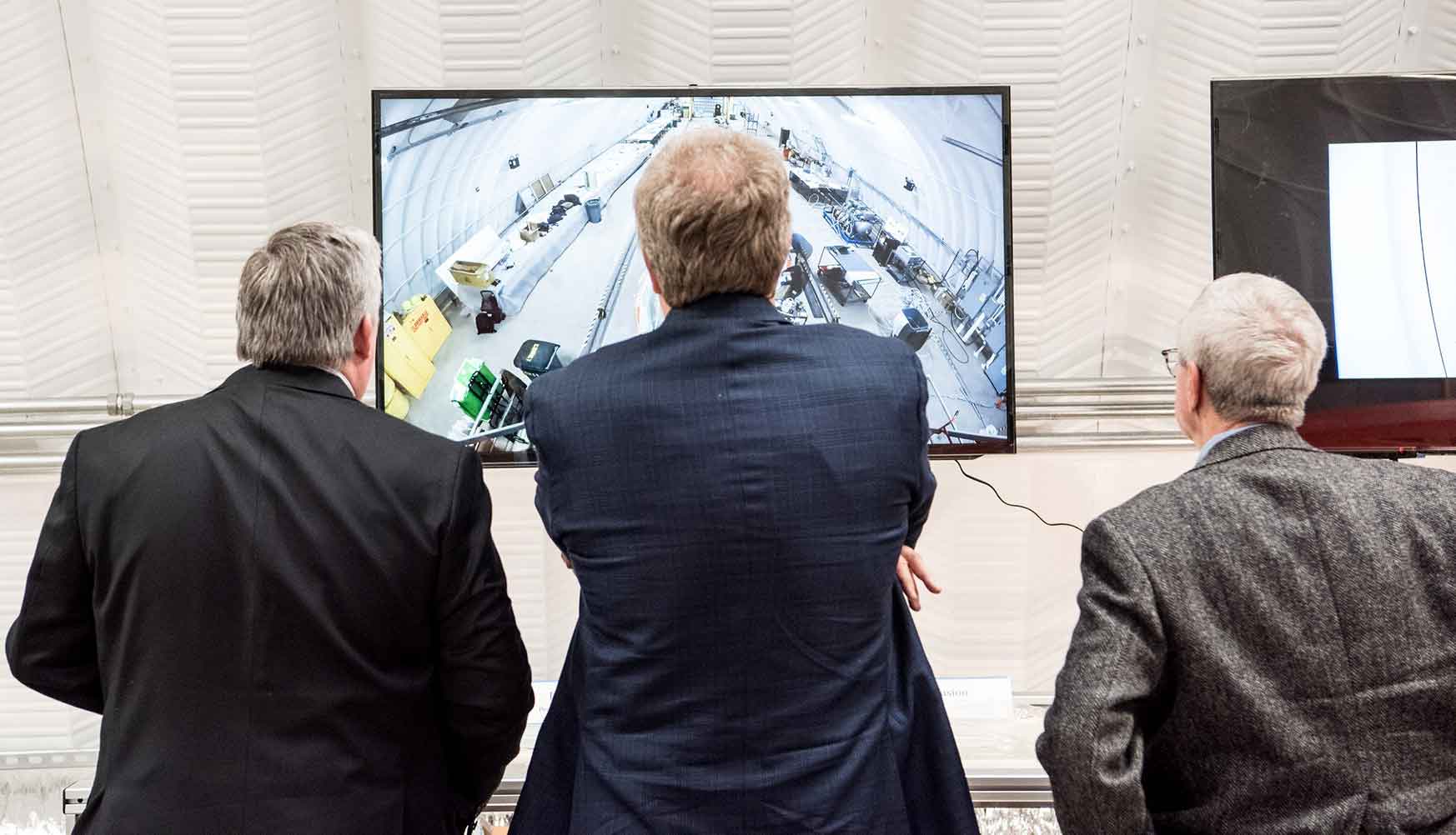 A photo of three men watching a television screen showing a time lapse video of the construction of a wind turbine blade. 