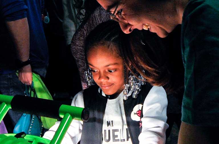 Photo of a young girl and a woman looking at a science experiment involving light emitted by a large, green lamp.