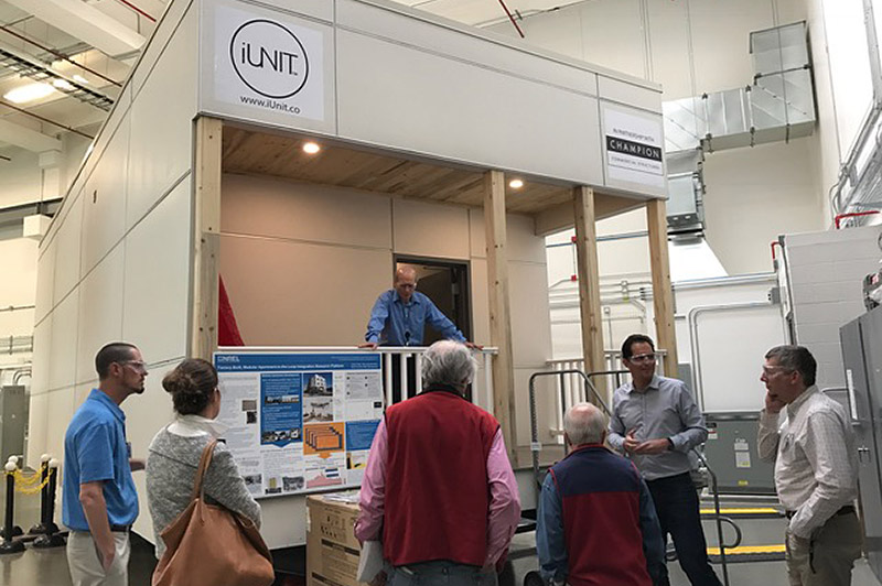 A photo of a group of people listening to a man speak inside a laboratory while another man stands on the balcony of a prototype modular studio apartment.