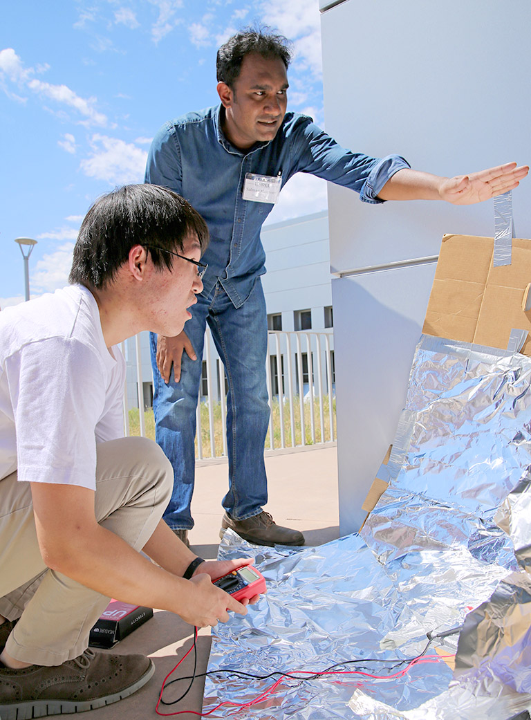 Two students, one standing, one crouching, examine a simple reflecting dish, made out of aluminum foil and cardboard. The dish is concentrating sunlight onto a solar cell, which one of the students is monitoring with a voltmeter.