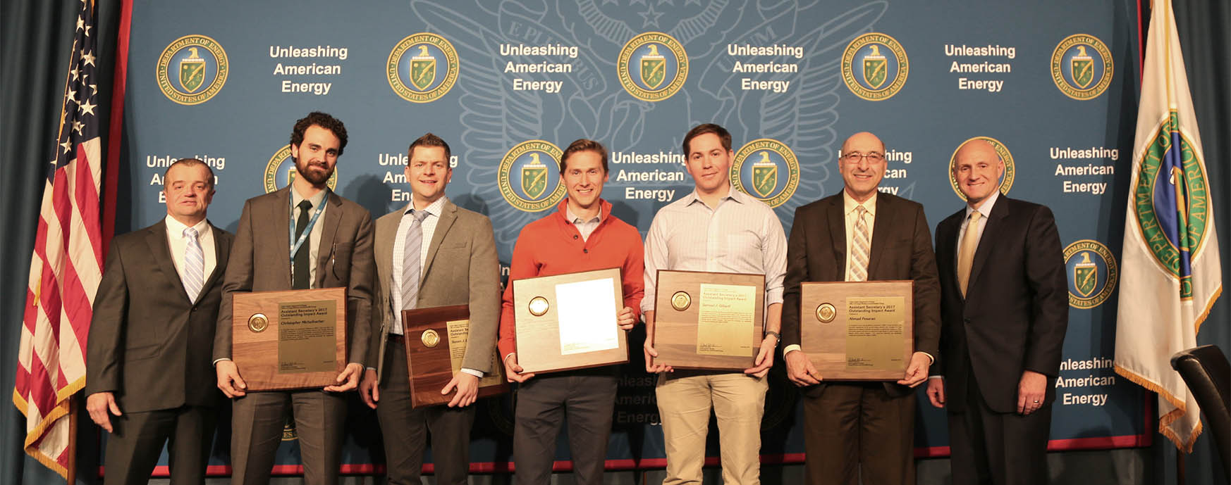 Men stand on a stage holding award plaques with flags and signage behind them.