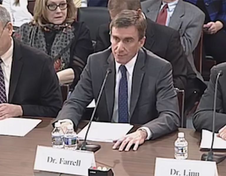 Photo of men seated at table with microphones in front of them and audience members behind them.