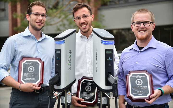 Three men hold awards and stand next to Electric Vehicle charging stations