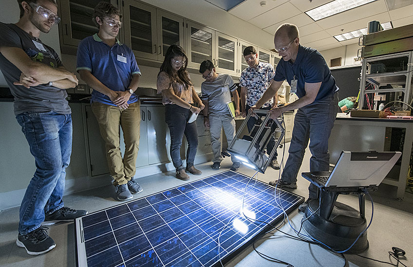 A group of students cluster around a PV module on the floor as an NREL scientist shines a light-camera combo on it, displaying the result on a nearby laptop. The goal is to visualize hidden cracks and defects in the panel.