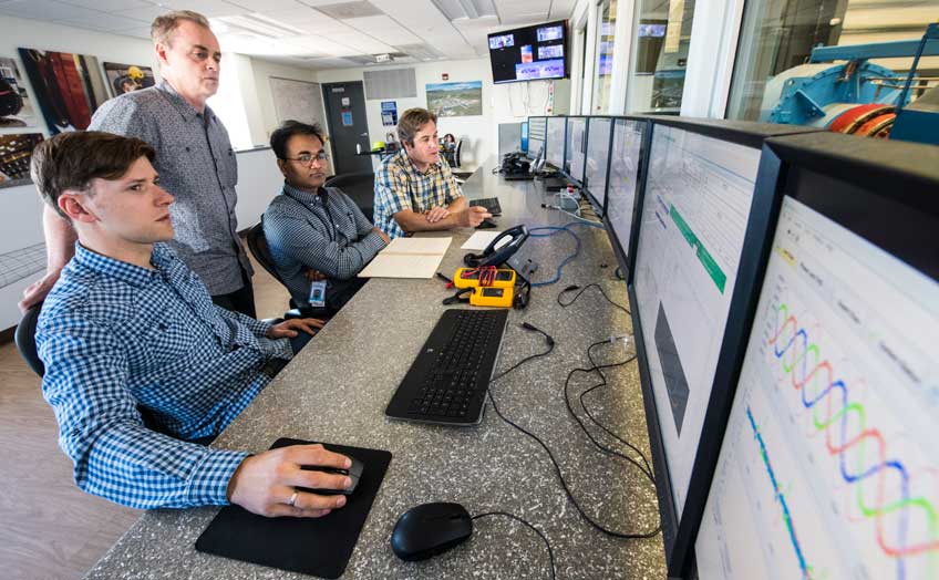 Four men (one standing, three sitting) look at rows of computer monitors.