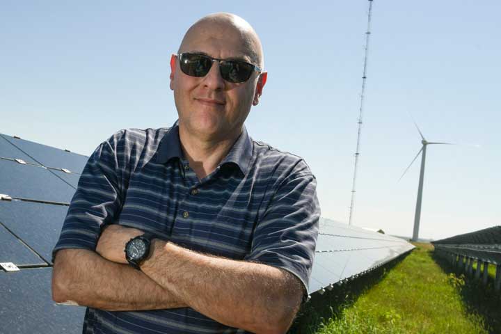 A man stands outside in front of a row of solar panels and near a wind turbine.