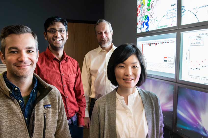 One female and three male researchers standing next to computer screens with computational modeling graphics on each screen.