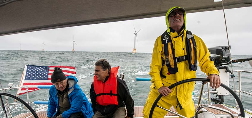 Two men sit and the captain stands, steering the boat. Behind them is an American flag on the boat and several wind turbines stand tall above the water.