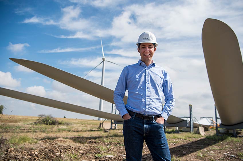 Man smiles in hardhat in front of wind turbine and three turbine blades.