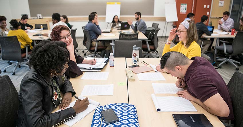 Photo of four students sit at a table laughing and writing down ideas as they work together on the mini-hackathon challenge held at NREL.