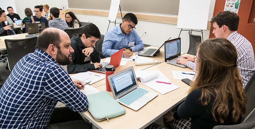 Photo of NREL engineer Tony Fontanini talking to a group of four students while they work on their laptops and write down ideas.