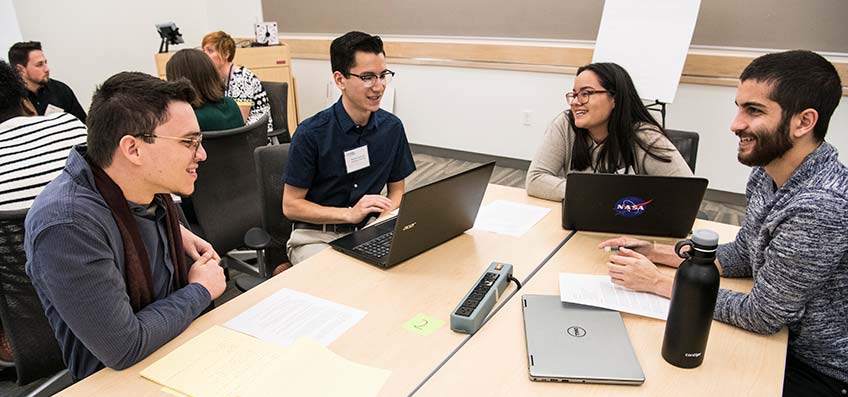 Photo of four students smile as they discuss ideas for the mini-hackathon challenge at NREL.