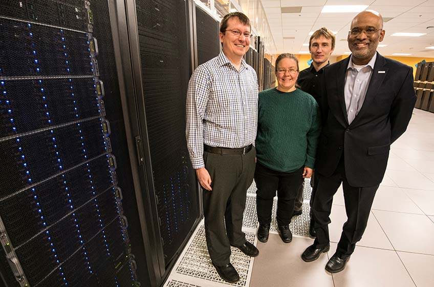 Four people stand next to a supercomputer.