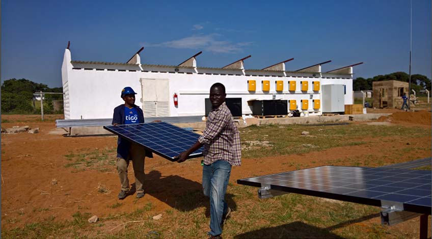 Two men carrying a solar panel in front of a white building.