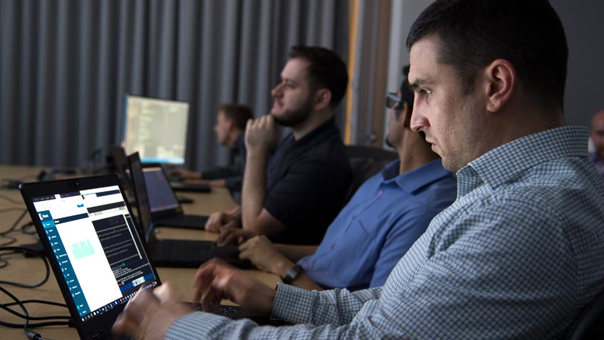 A man in the foreground types on a computer pad while several others sit nearby.