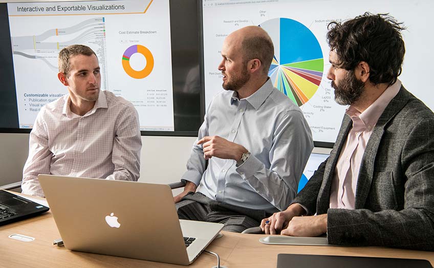 Three men talking in front of large computer screens.