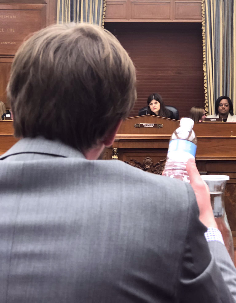Photo of a man holding a plastic water bottle and speaking to a panel of U.S. Representatives.