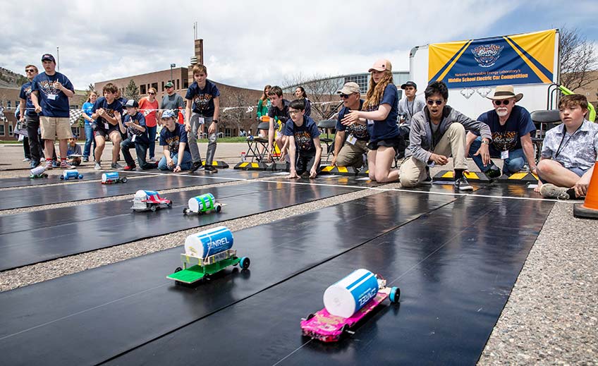 Model cars race down a track as a group of students cheer at a model electric car competition.