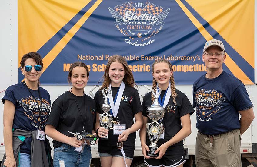 Five people smile as they hold their 1st and 3rd place trophies at the awards ceremony of the middle school electric car competitions.