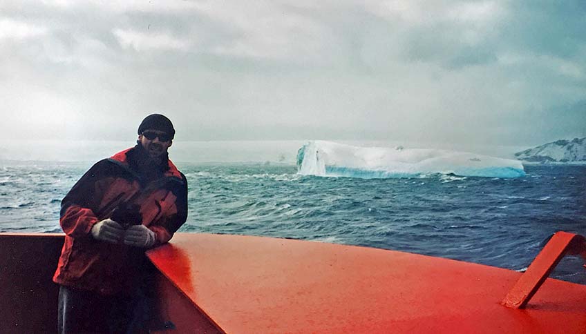 A man in a heavy coat onboard a ship on the ocean, with icebergs in the background