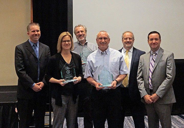 Six people standing as a group and smiling. Three of them are holding awards.