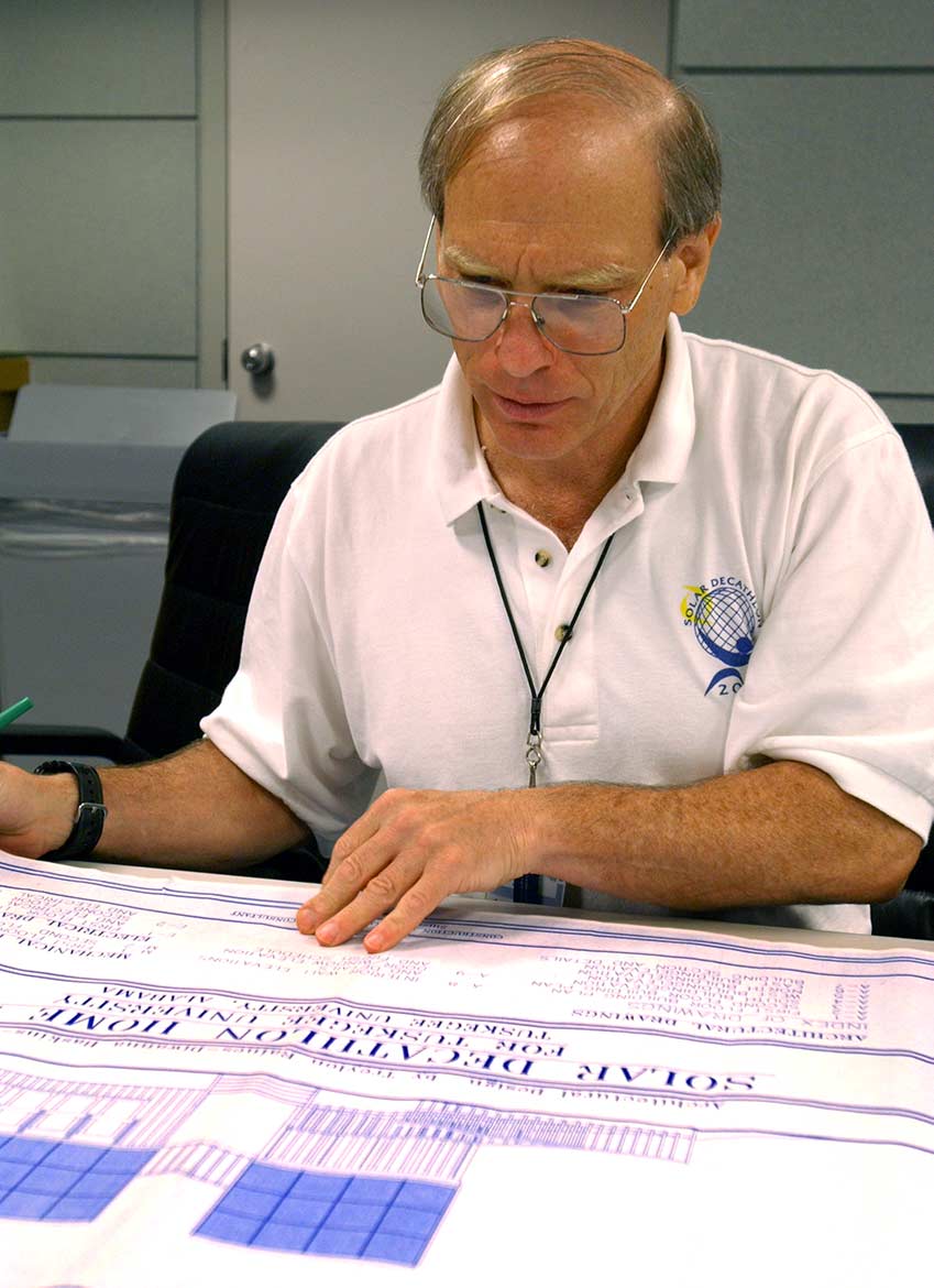 Ron Judkoff sits in a white collared shirt with a Solar Decathlon logo, reviewing a document.