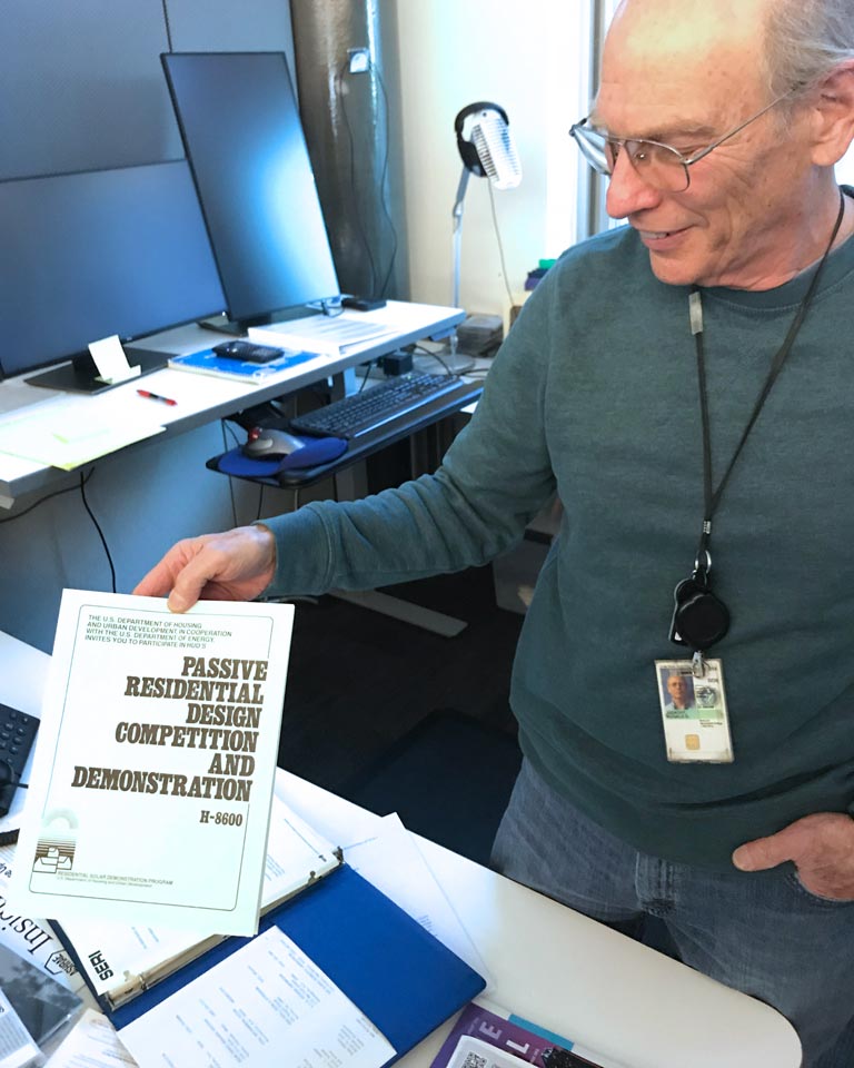 Ron Judkoff stands in an office holding a copy of a manual titled "Passive Residential Design Competition and Demonstration."