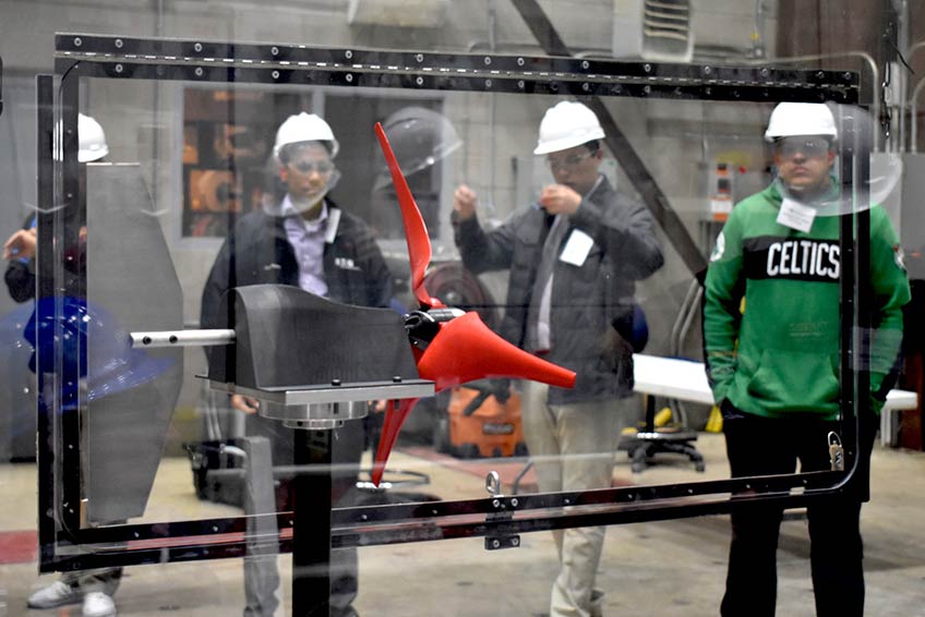 Five male students wearing safety glasses, ear plugs, and hard hats stand behind protective glass that houses a black model sized wind turbine with red blades.