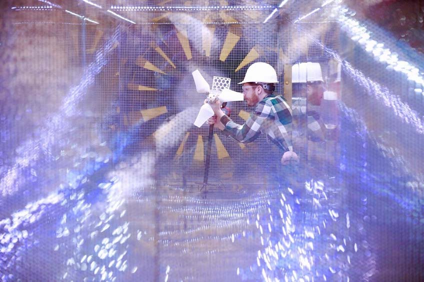 A young man with a red beard and hard hat adjusts a small-scale wind turbine inside a wind tunnel.