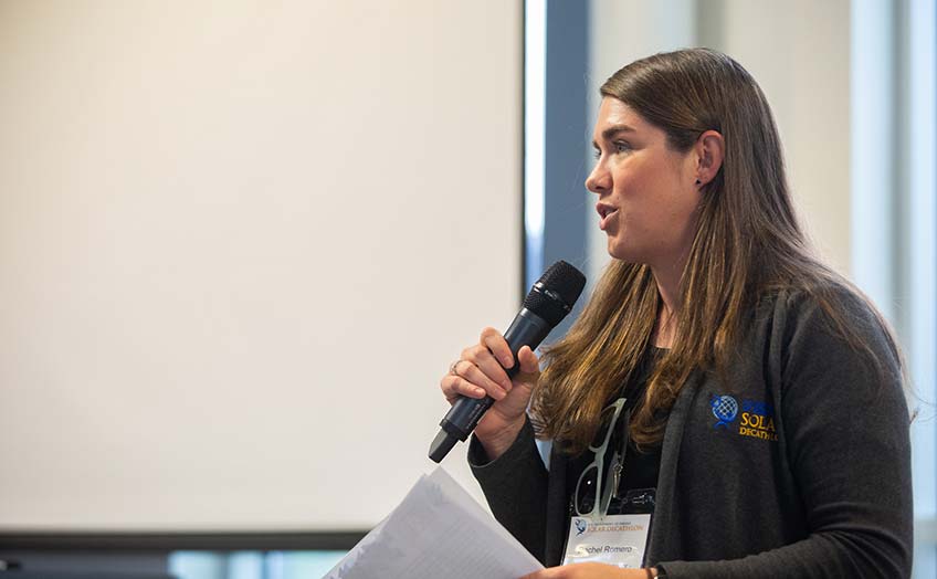  Rachel Romero speaks into a microphone in a jacket with a Solar Decathlon logo.