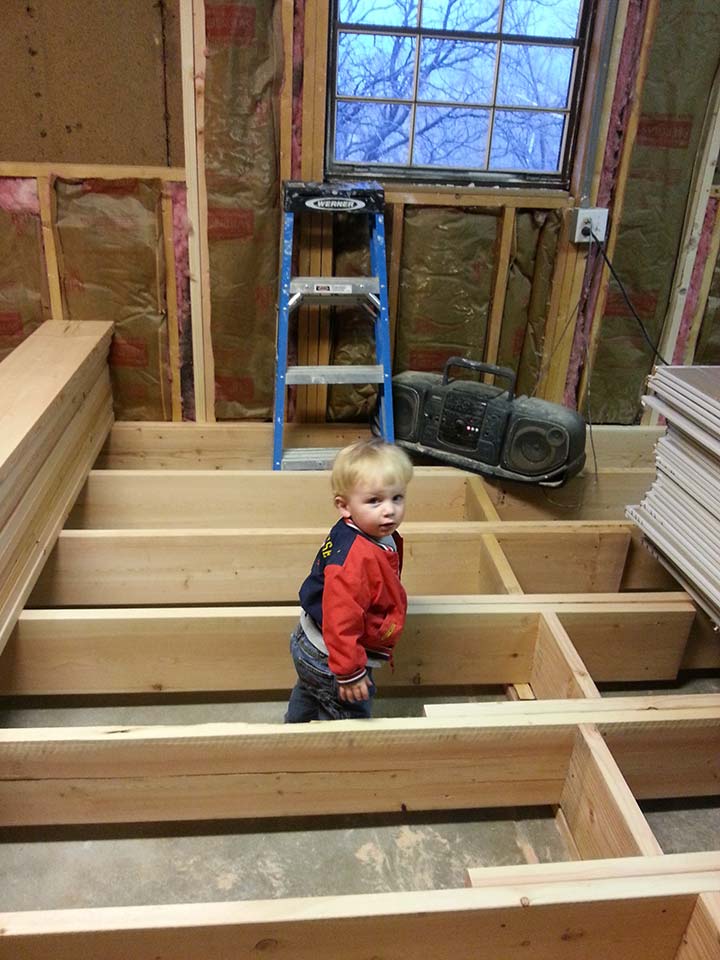 A toddler stands in an unfinished attic next to a ladder and a stack of HVAC filters.