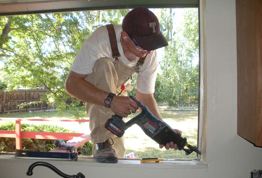 Booten wears safety googles, overalls and a baseball cap while crouching at work in a windowsill.