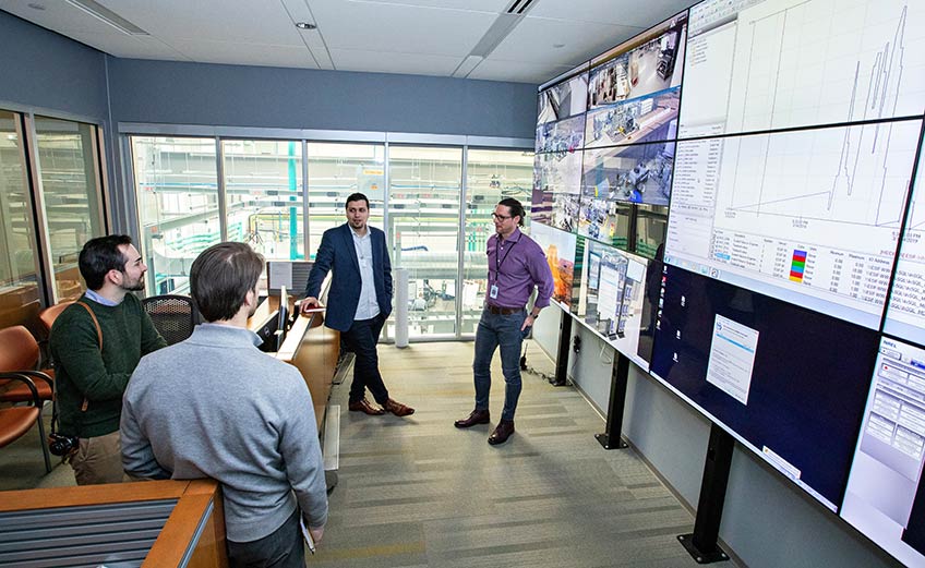 Photo of 4 men in coversation next to a wall of screens with multiple graph projections