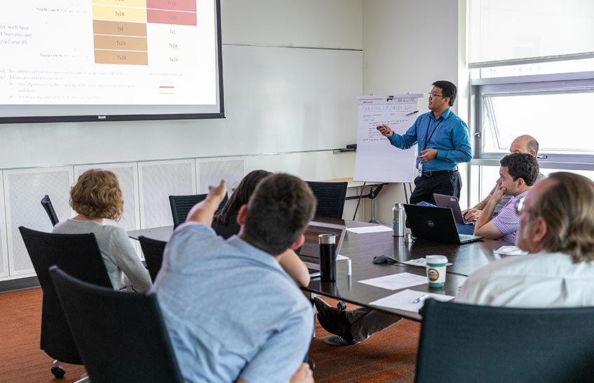 A man presents a PowerPoint slide in front of a group of people in a conference room.